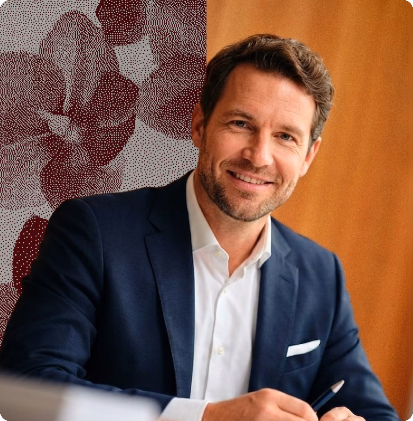 Homme souriant en veste bleue et chemise blanche assis, tenant un stylo avec un fond marron et motif.