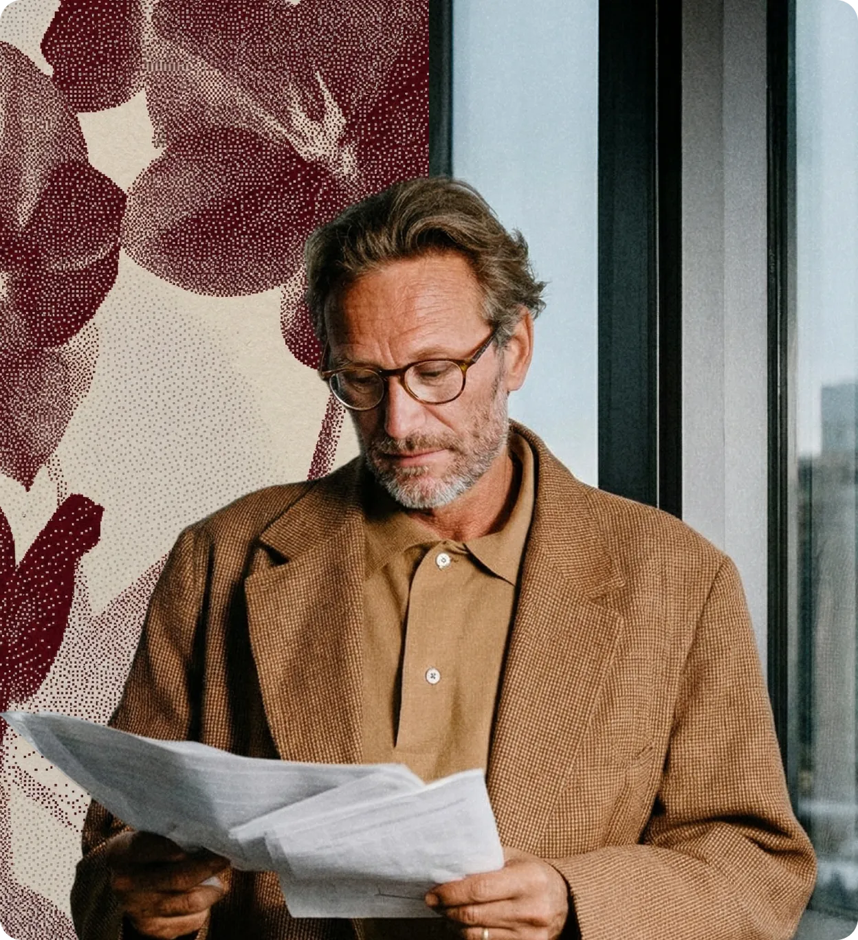 Homme à lunettes lisant des documents, portant un manteau marron devant un mur orné de feuilles rouges.