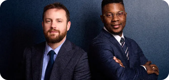 Deux hommes d'affaires posant devant un mur bleu, l'un avec barbe, l'autre bras croisés, en costume.
