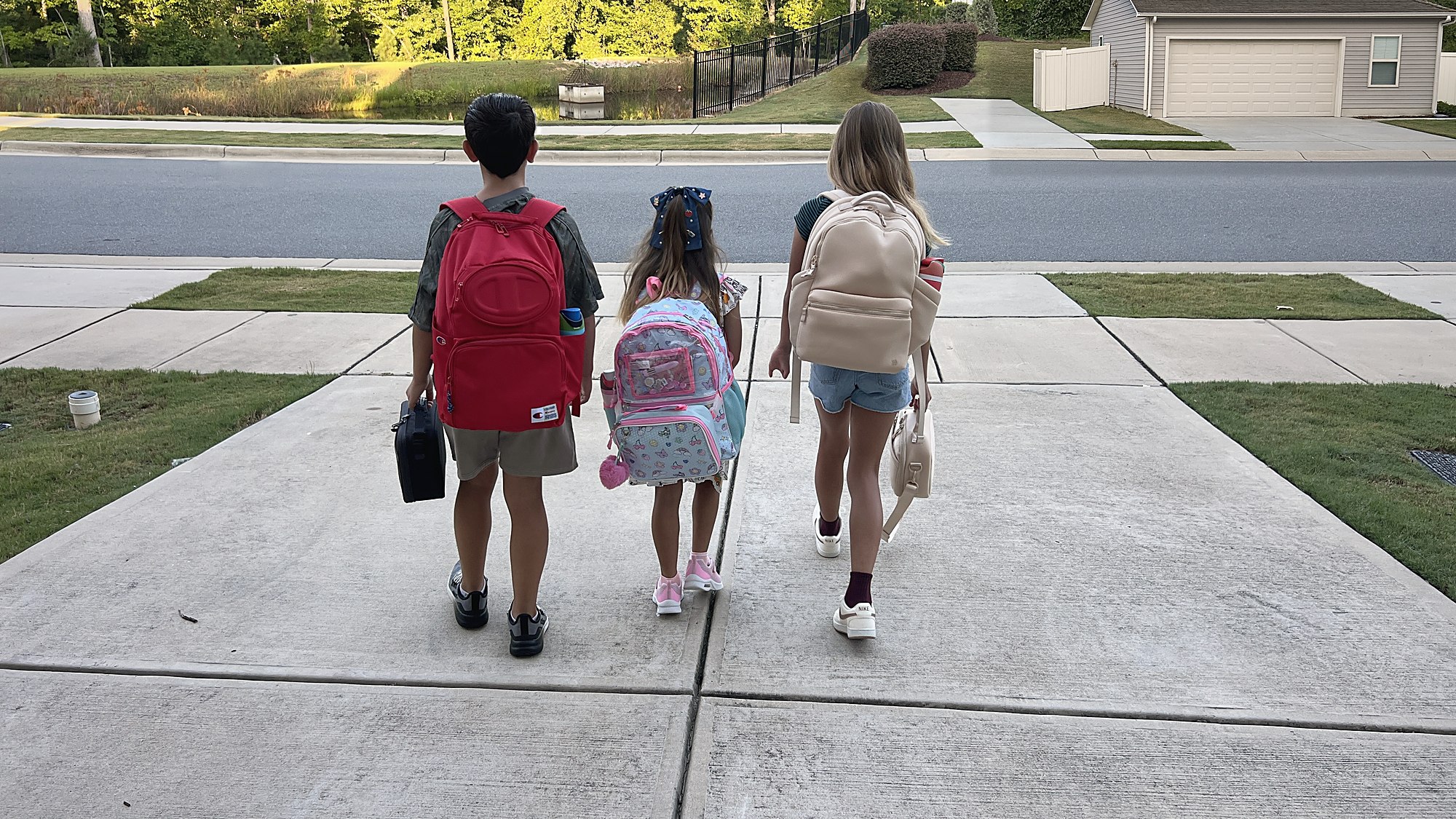 Three kids with backpacks walking down the driveway on the first day of school