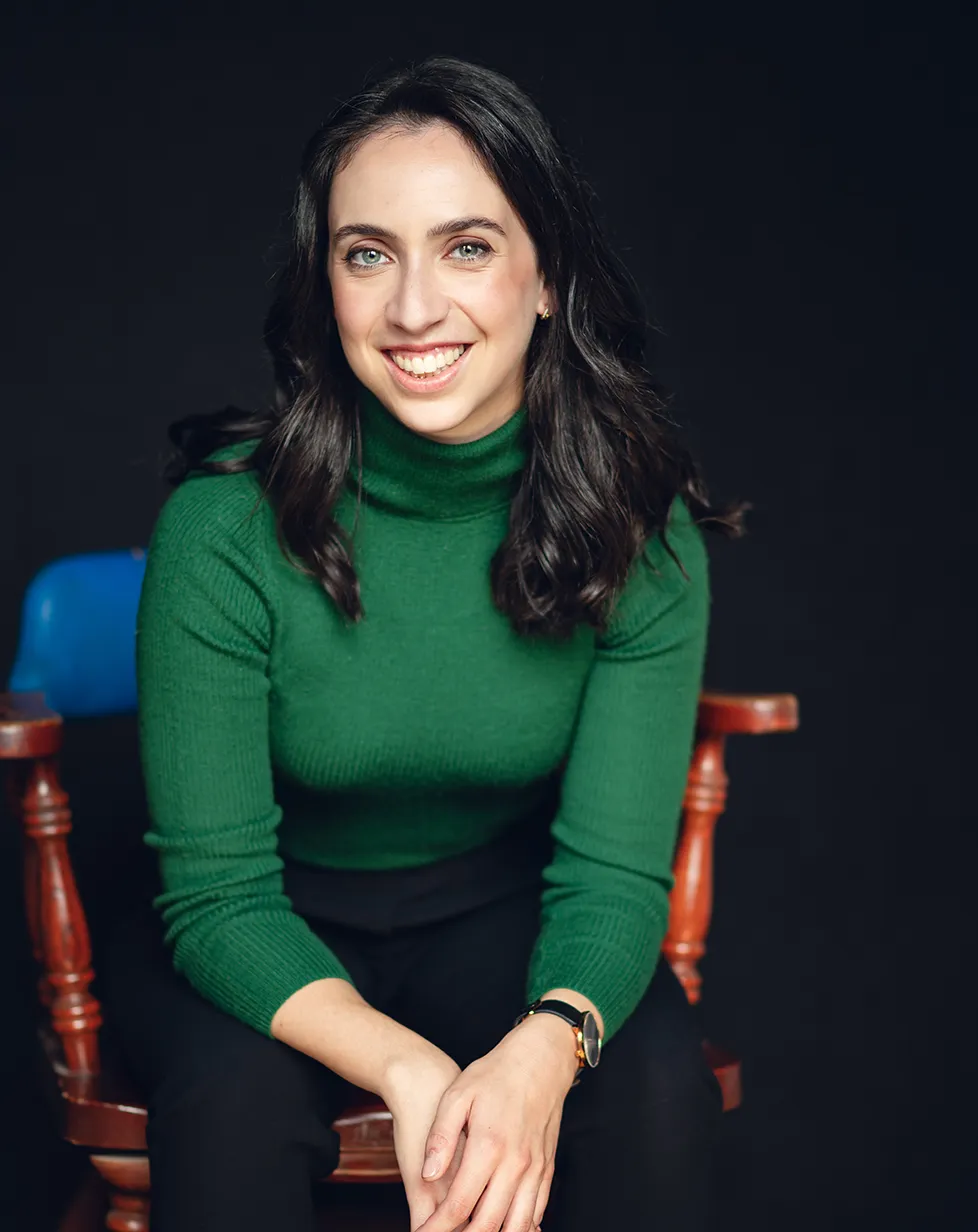Martine est assise face à la caméra dans un fauteuil orange et blanc aux finitions en bois. Vêtue d’un chandail vert, elle affiche une expression accueillante qui semble dire : « Je suis prête à travailler avec vous. Faisons de la musique ensemble ! »