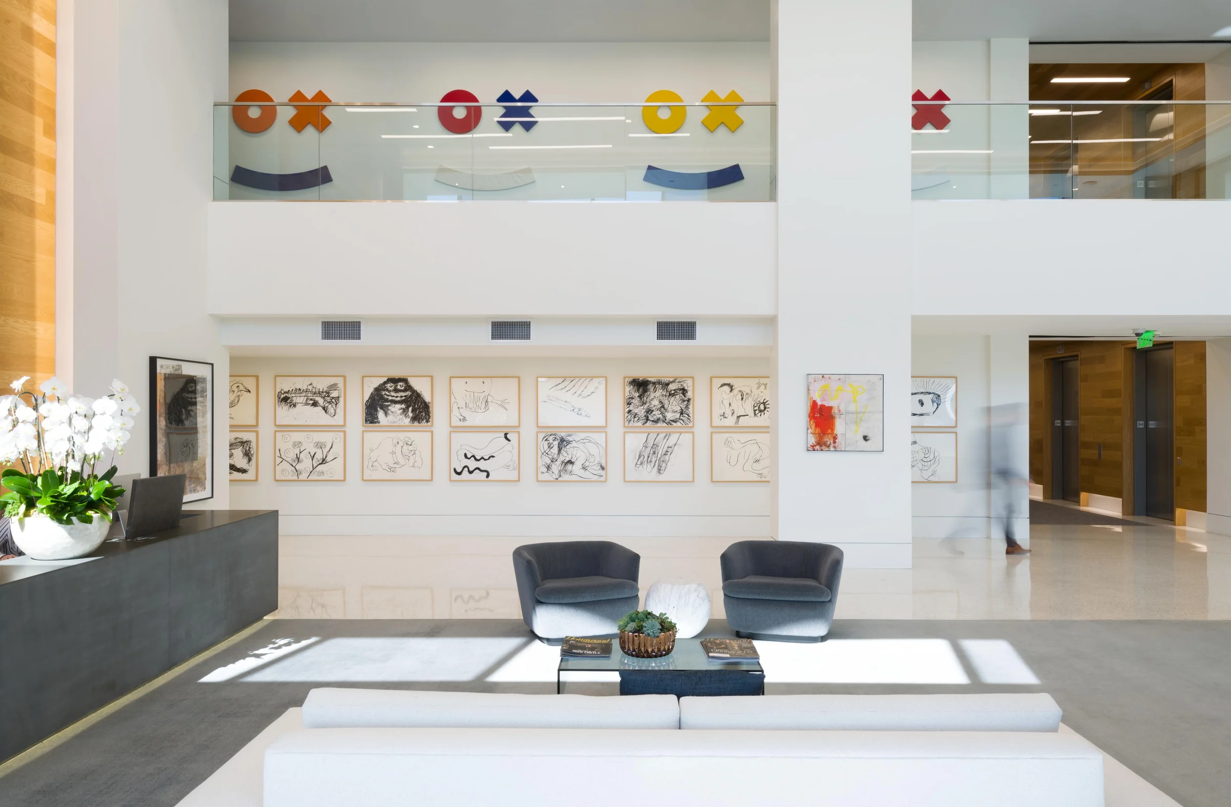Double-height corporate lobby with a grid of framed drawings and colorful wall sculptures on the mezzanine