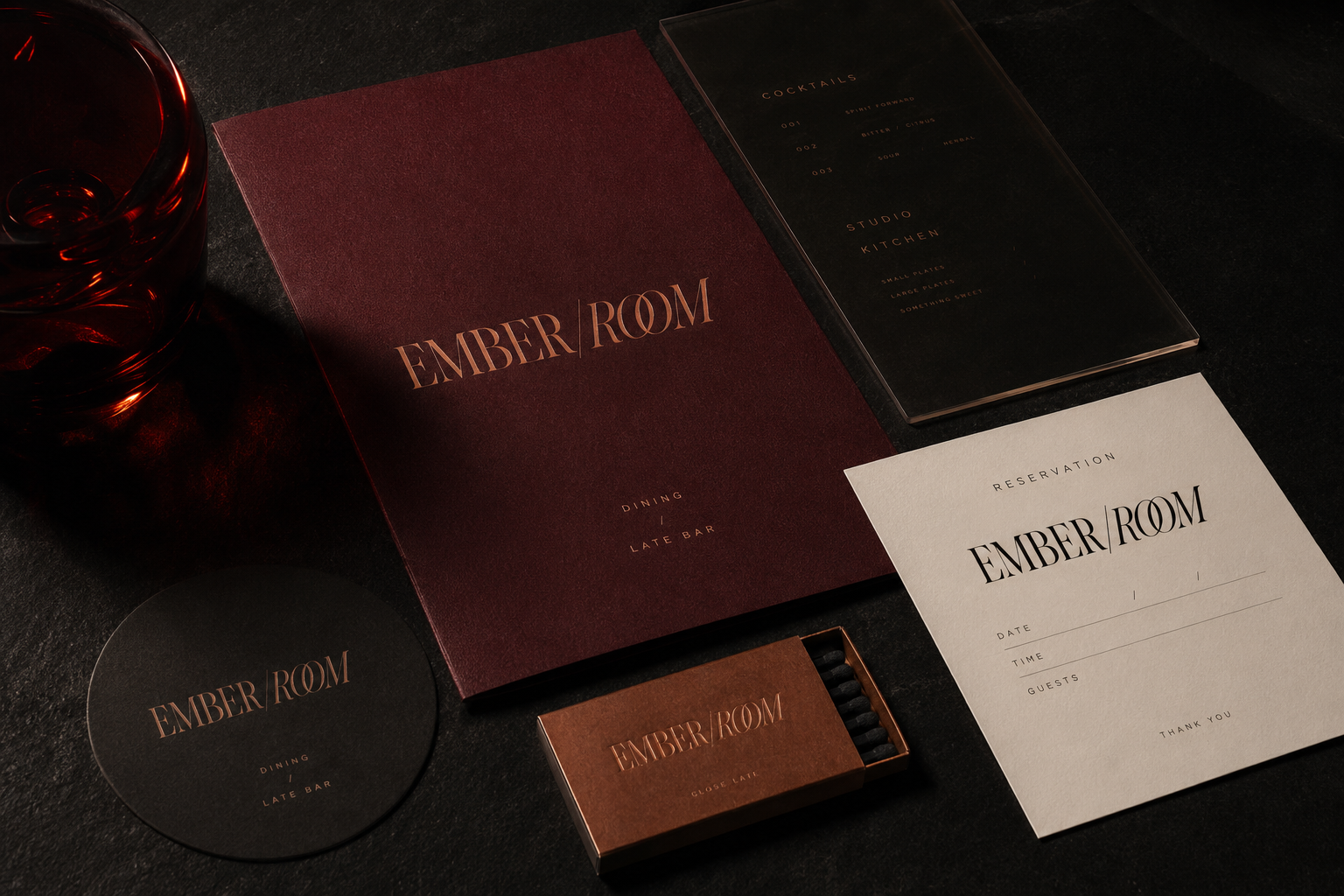 Table with Ember Room branded items: menus, coaster, reservation card, matchbox, and a red glass.