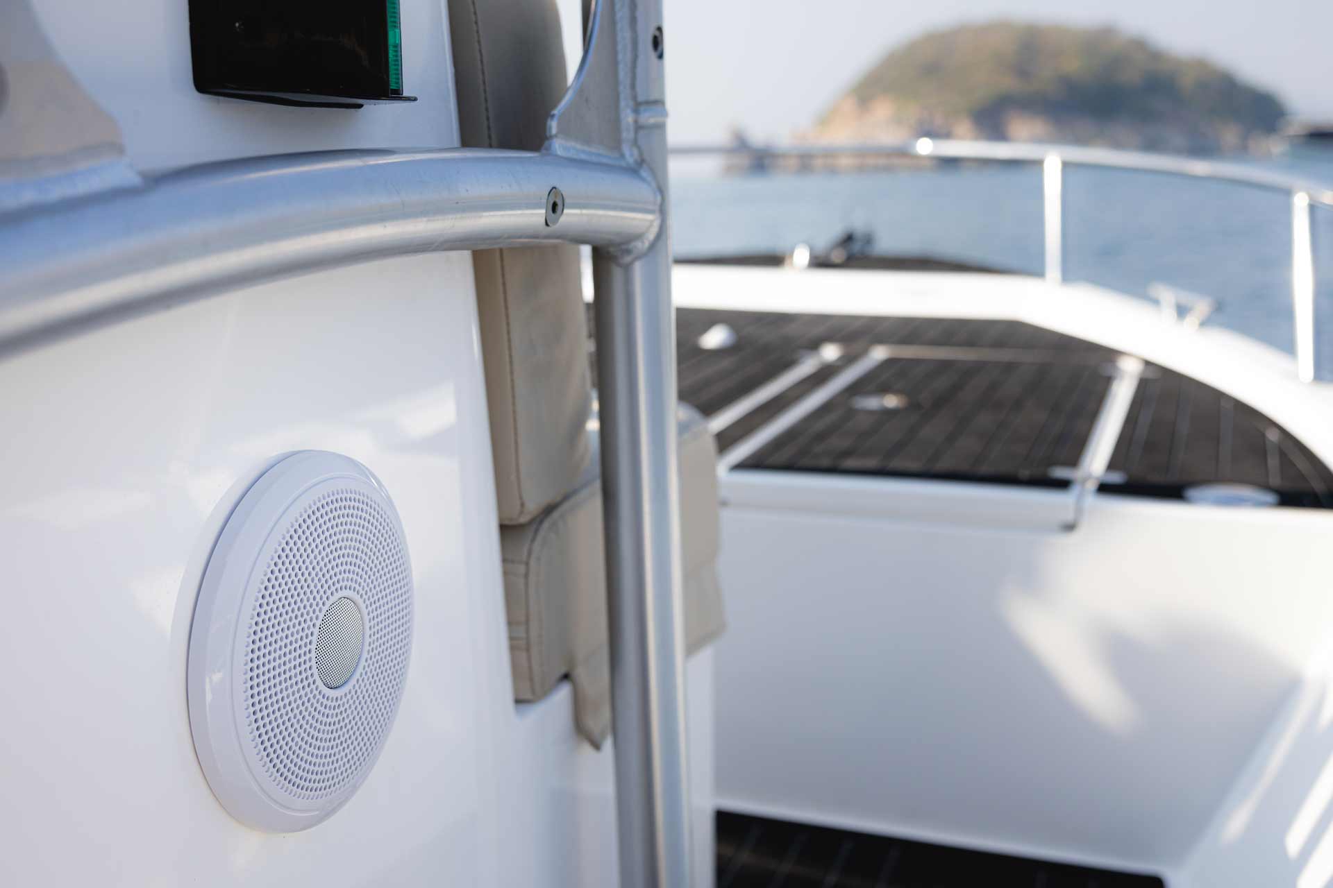 Close-up of a white marine speaker mounted on a boat railing with water and a small island in the background.