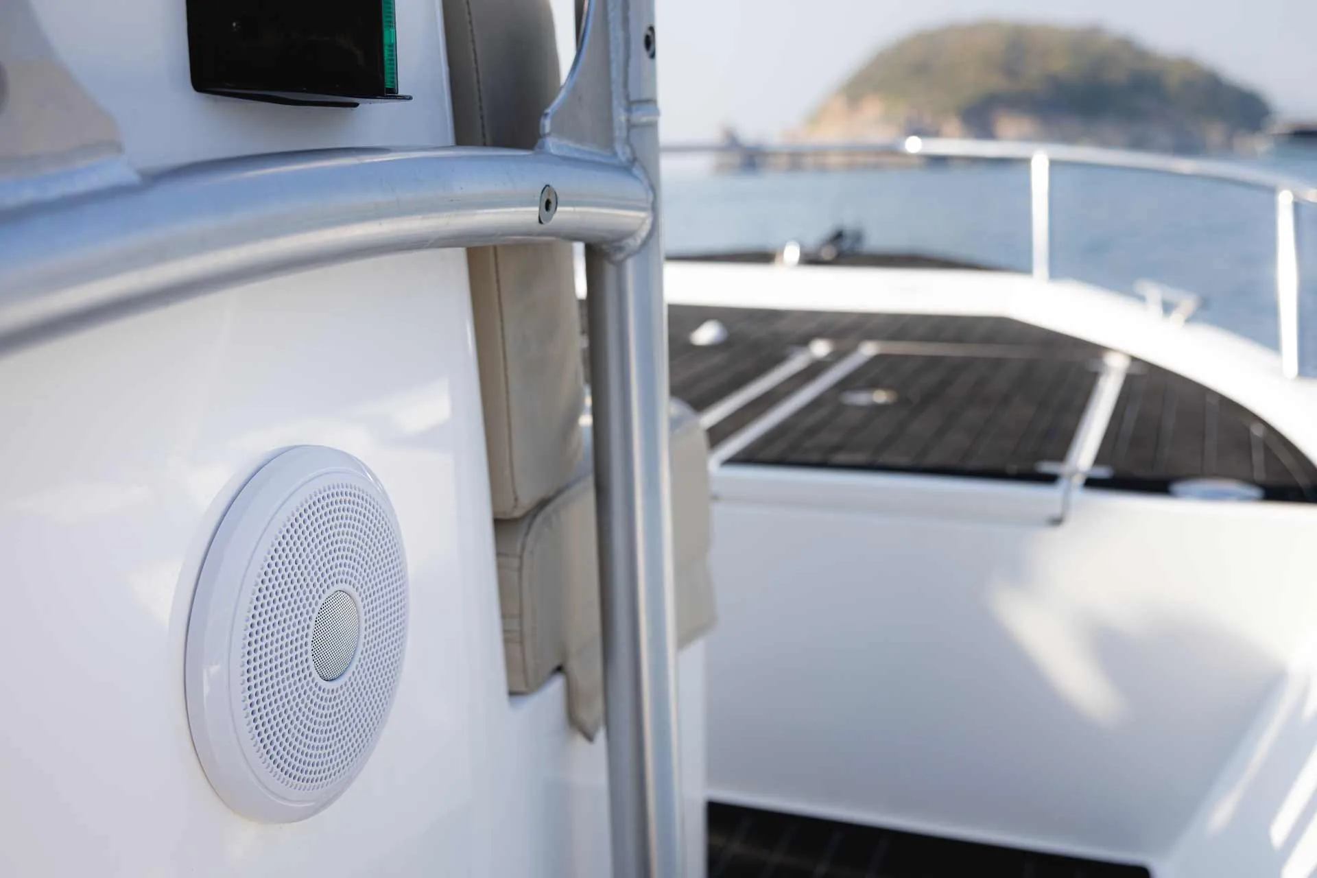 Close-up of a white marine speaker mounted on a boat railing with water and a small island in the background.