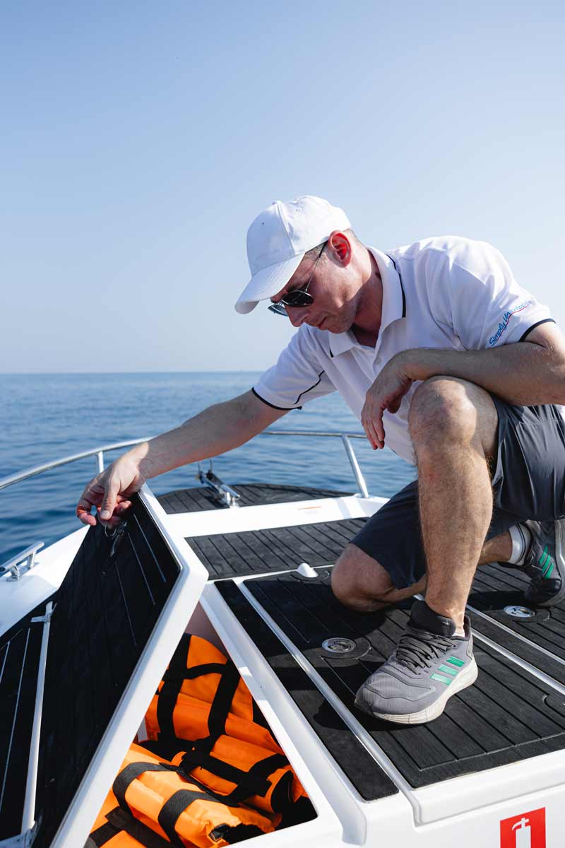 Man in a white cap and sunglasses opening a storage hatch on a boat revealing orange life jackets inside.