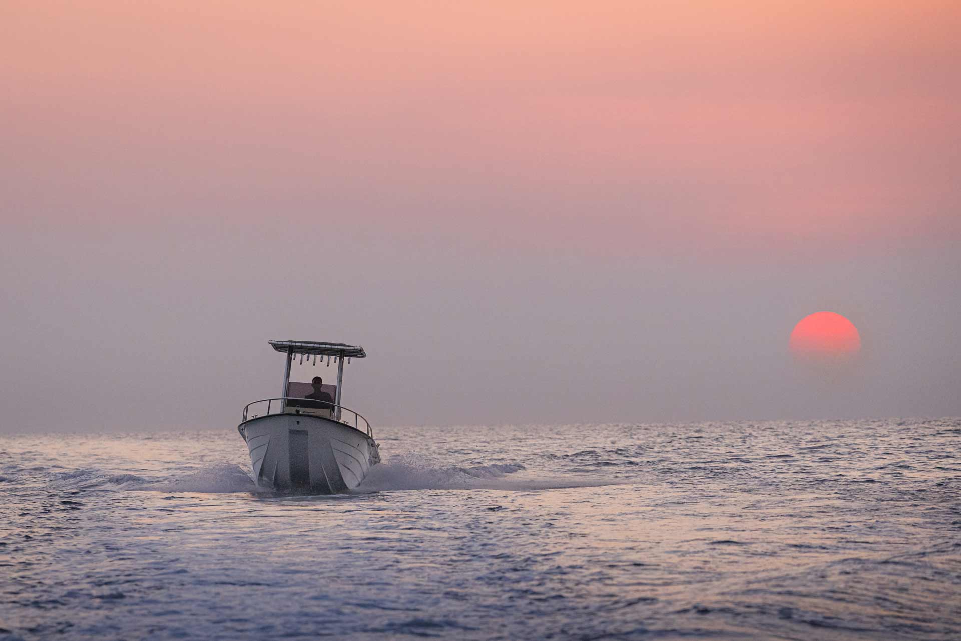 A boat with a person on board moving across calm water at sunset with a red sun near the horizon.