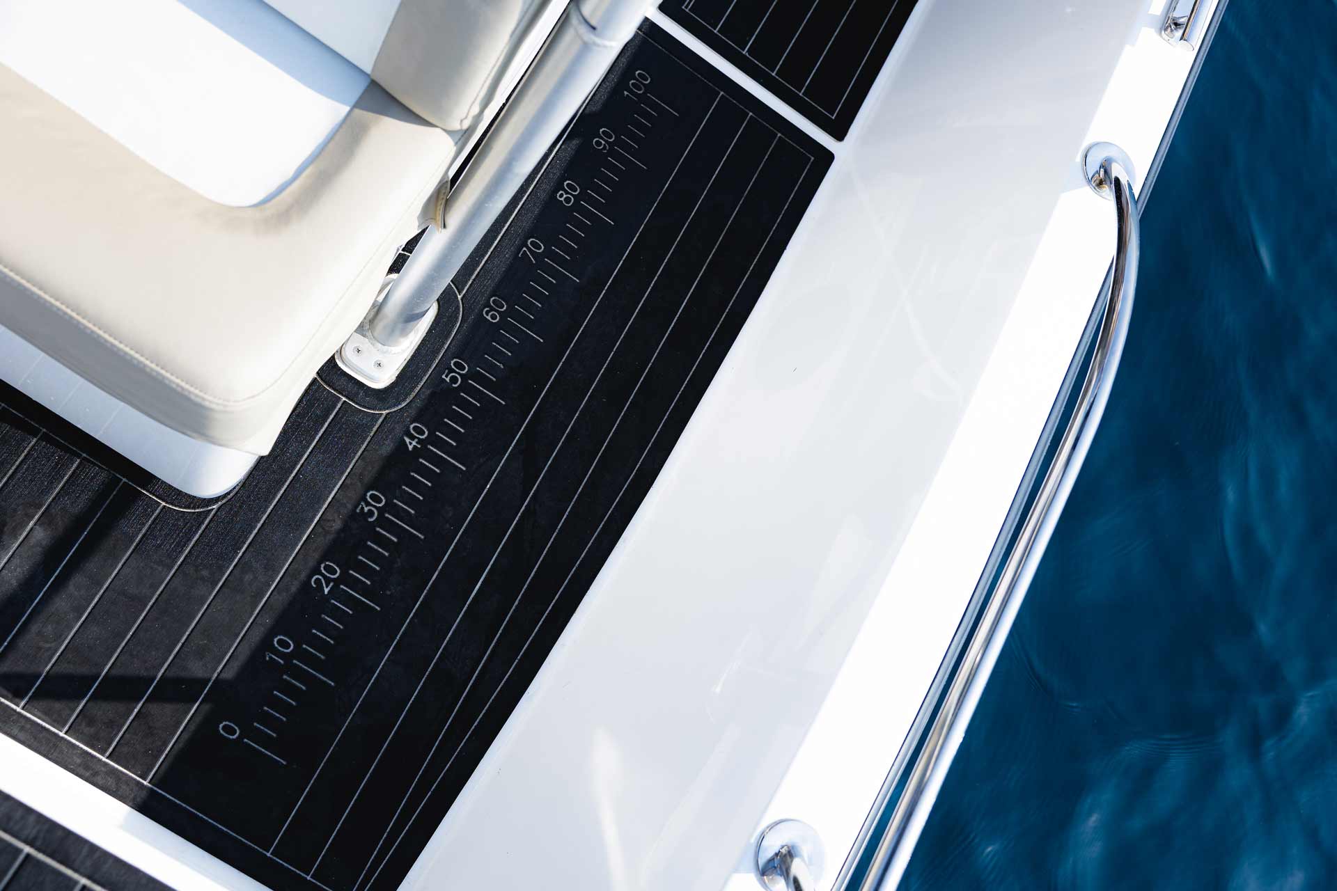 Close-up of a boat deck corner with black numbered measuring mat, white cushioned seating, and metal railing over blue water.
