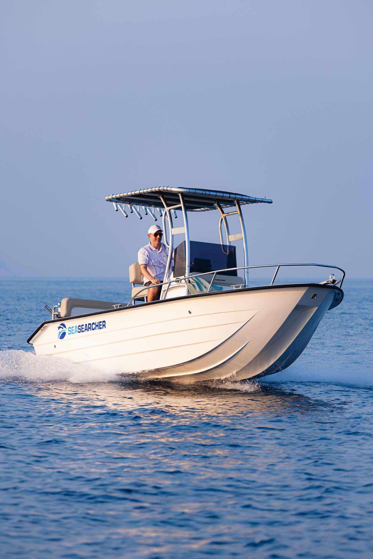Man wearing white cap and shirt steering a small white motorboat named SEASEARCHER on calm blue water.