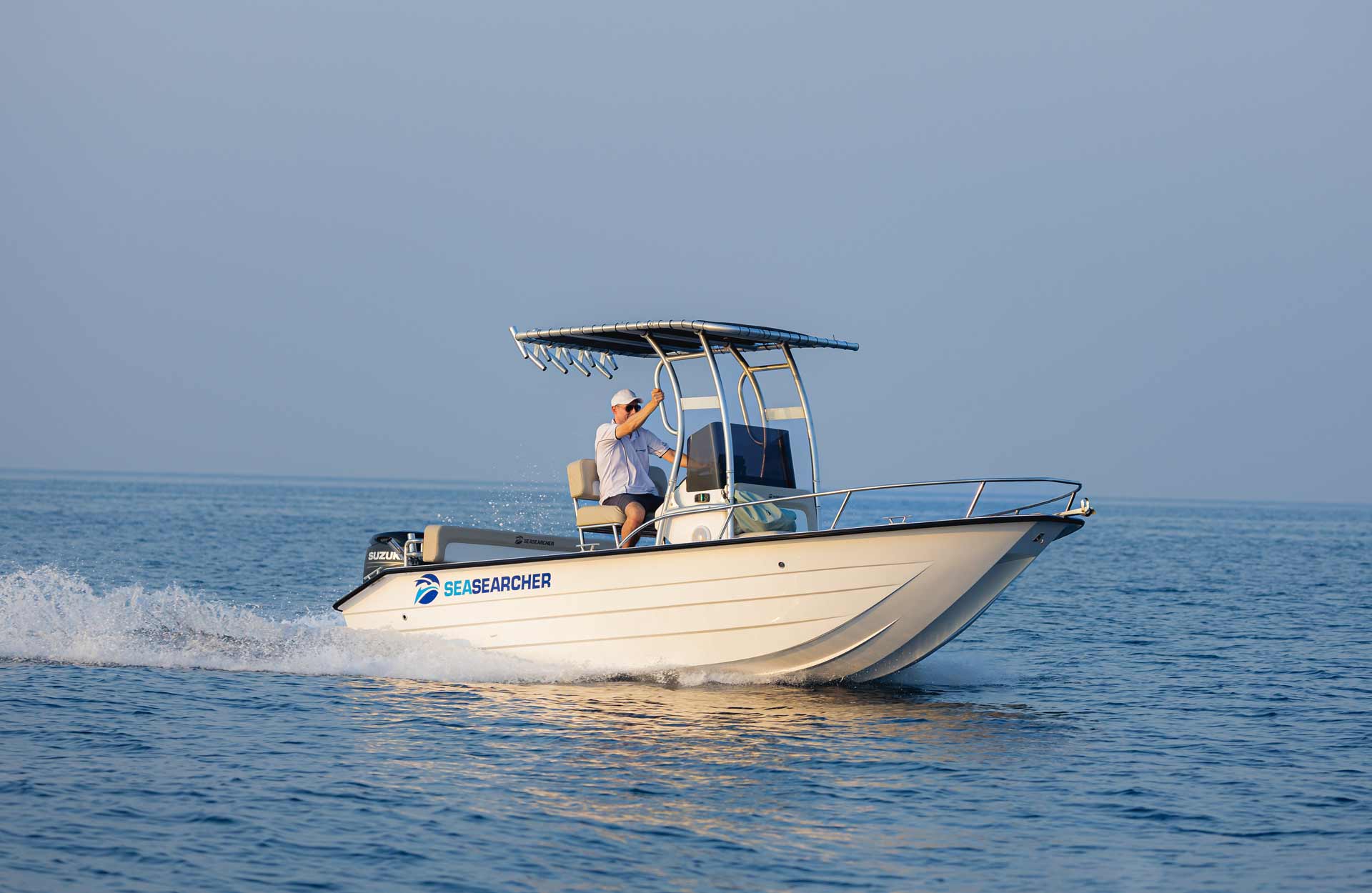 A man wearing a cap and sunglasses operates a white SeaSearcher motorboat on calm blue water with clear skies.