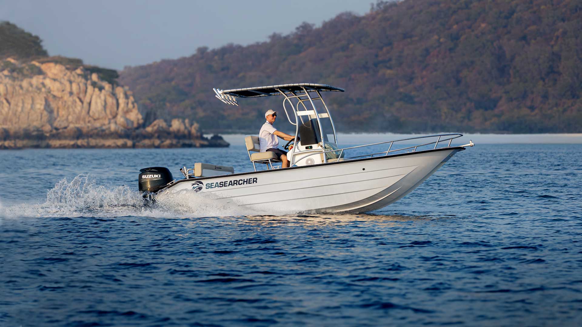 Man driving a white SeaSearcher motorboat on calm blue water with rocky and forested hills in the background.
