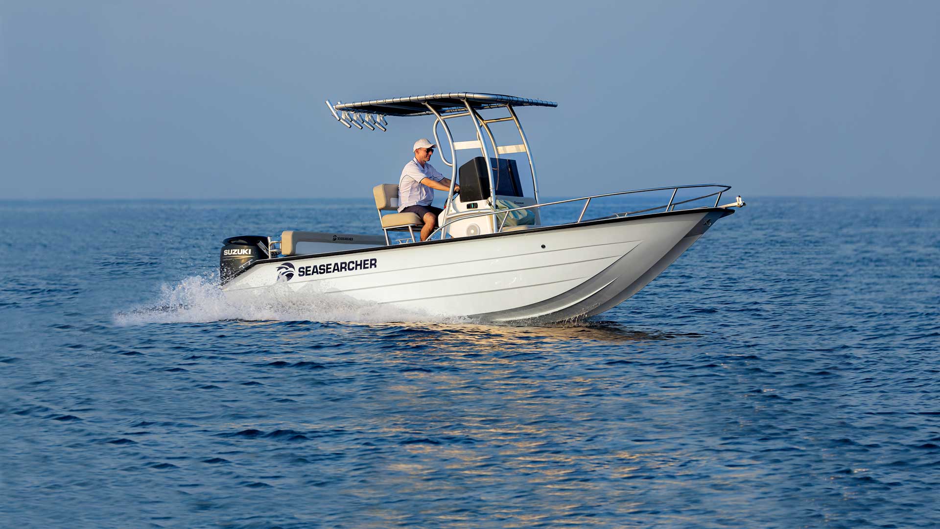 Man piloting a white SeaSearcher motorboat on calm blue water under clear sky.