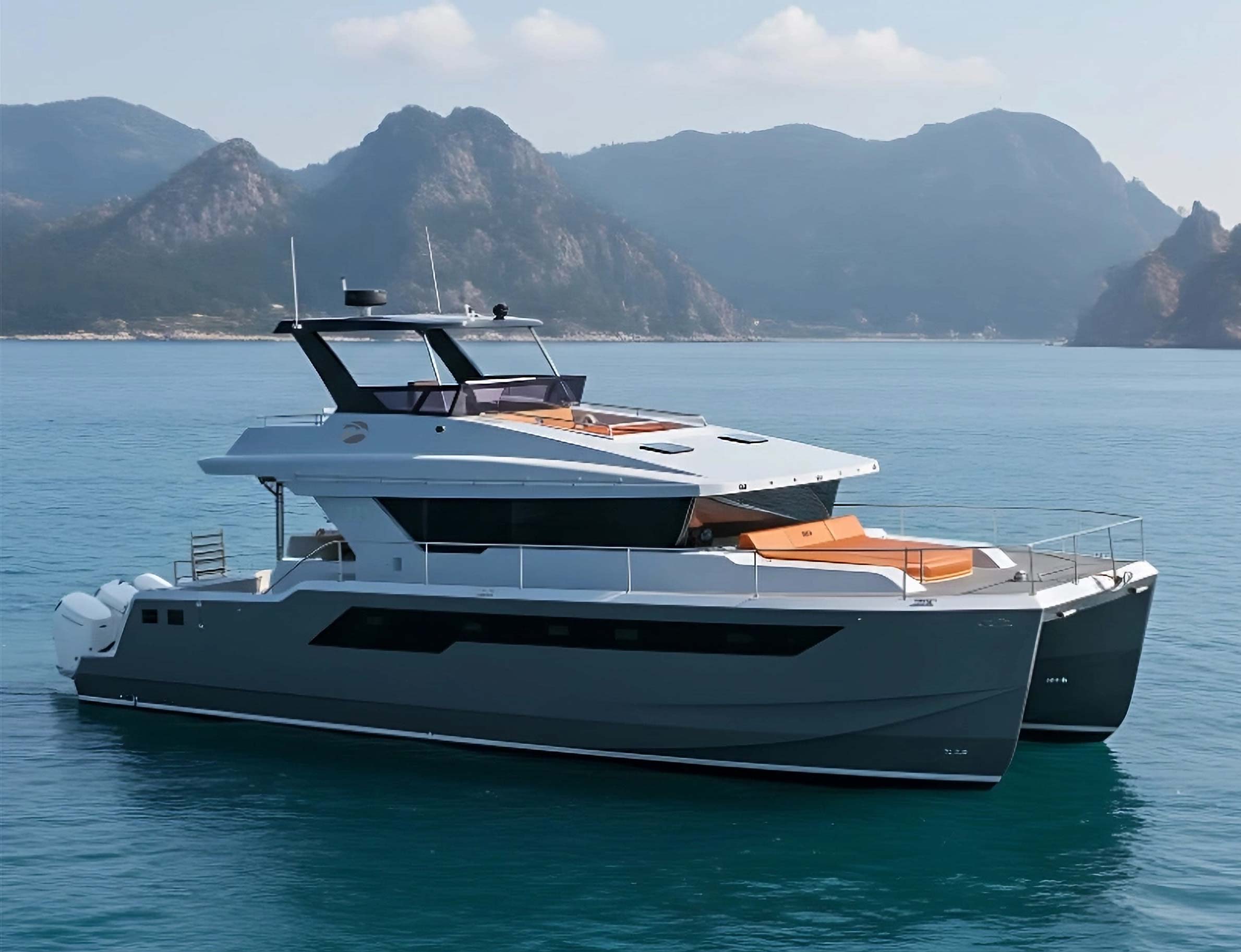 Modern catamaran yacht with orange cushions floating on calm blue water with mountain backdrop.