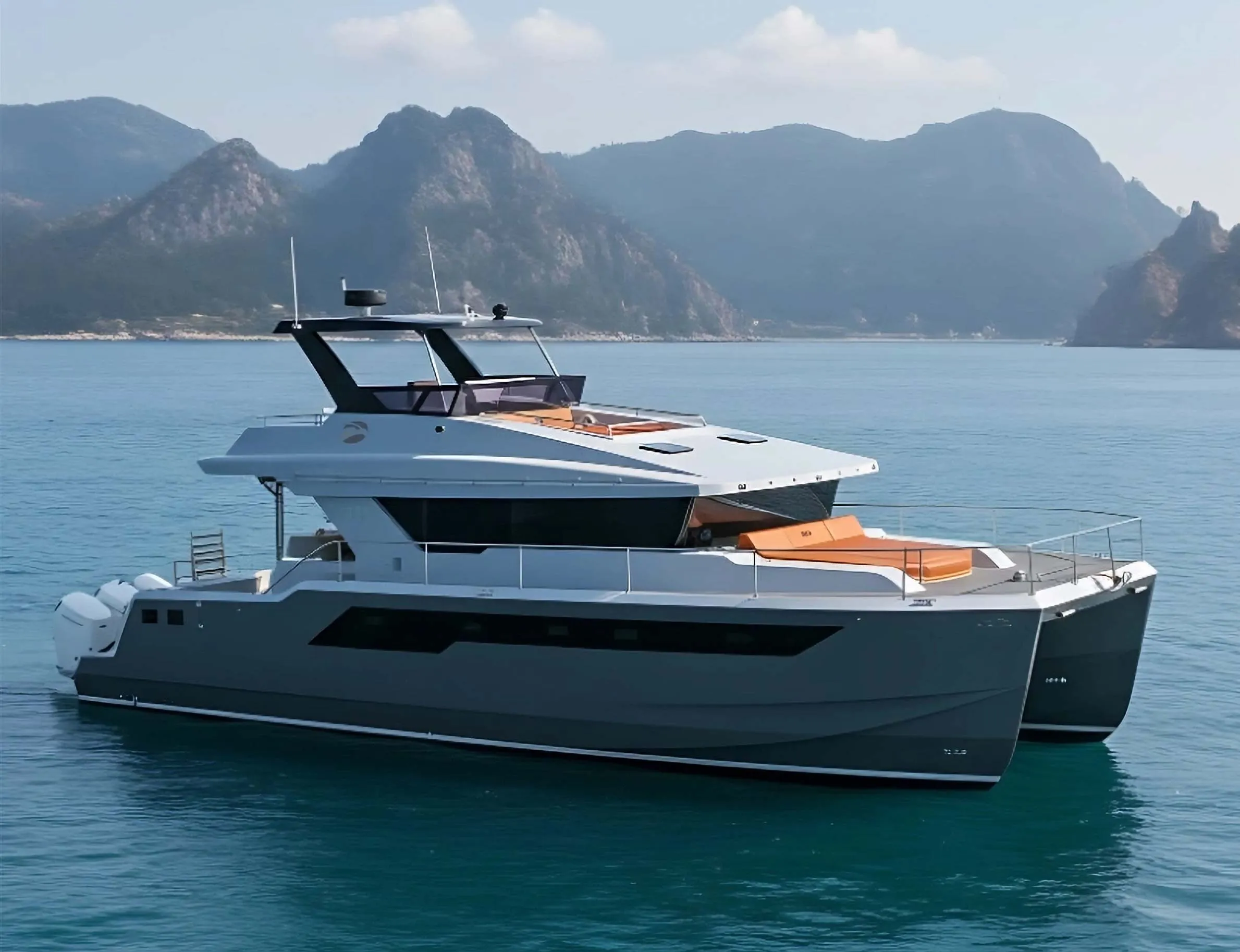 Modern catamaran yacht with orange cushions floating on calm blue water with mountain backdrop.