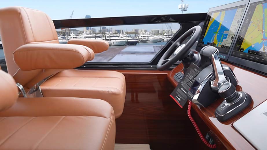 Interior of a boat cockpit showing two beige leather pilot seats, a steering wheel, and dual navigation screens displaying marine charts.