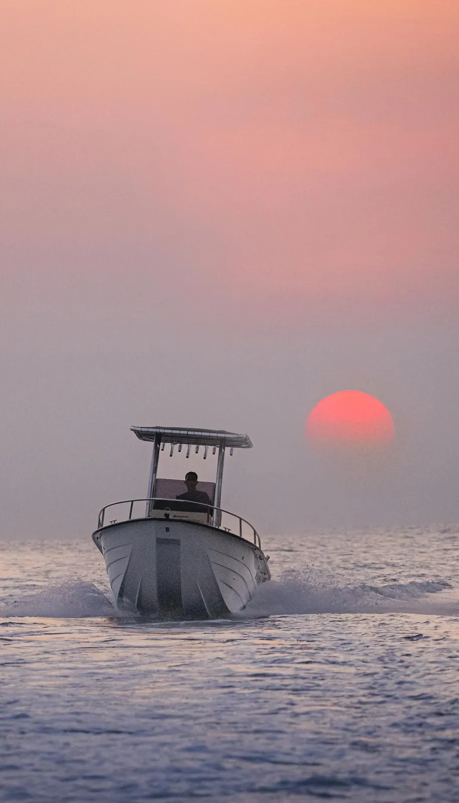 Boat with a person driving on ocean water at sunset with a large red sun near the horizon.