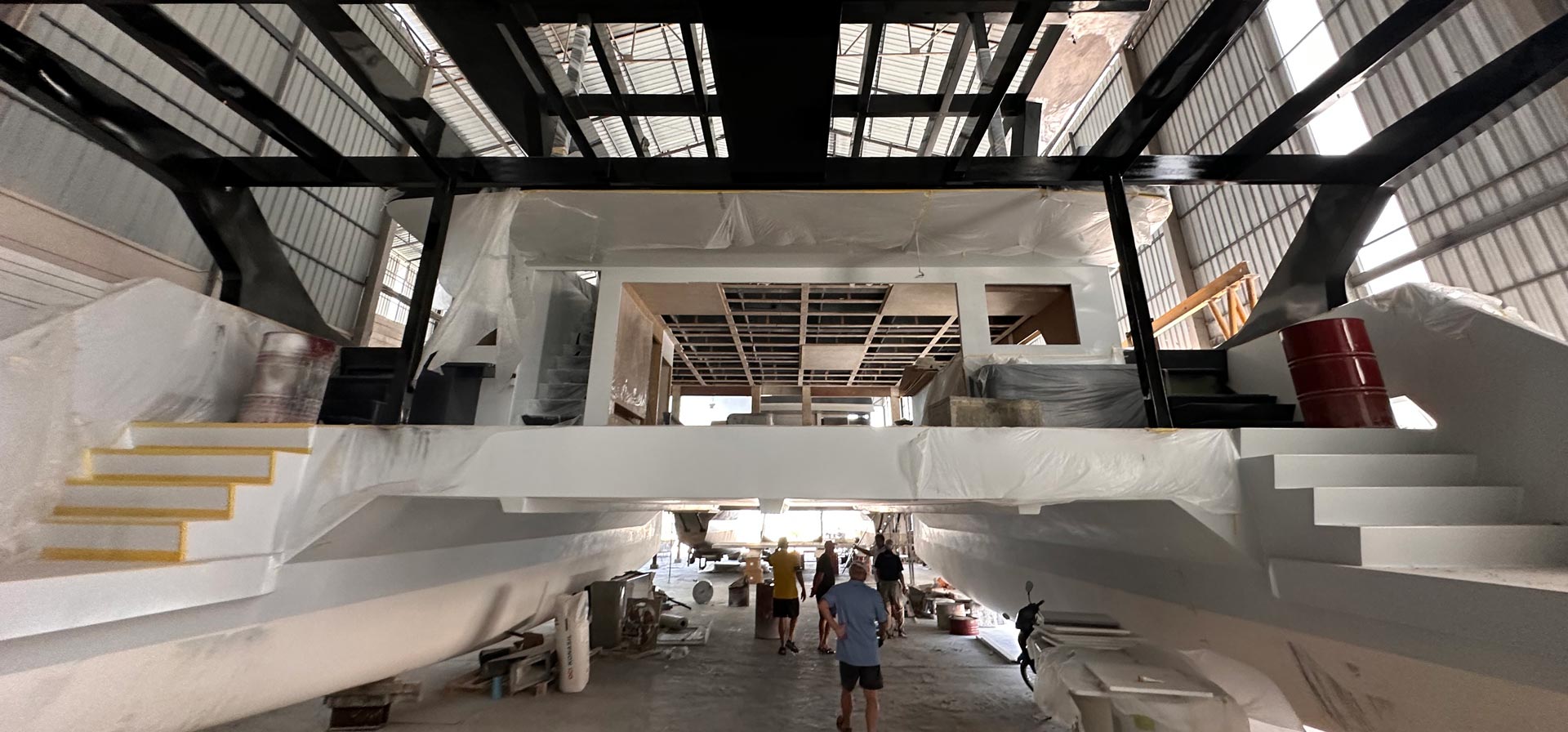 Construction phase of a large white catamaran boat inside a warehouse with workers walking underneath the raised deck.