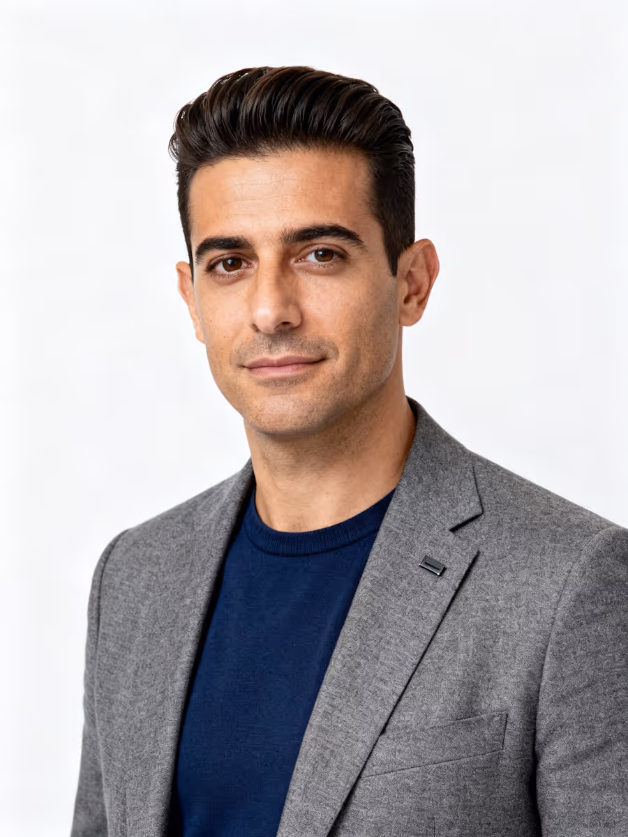 Portrait of a man with dark hair wearing a gray blazer and navy blue shirt against a white background.
