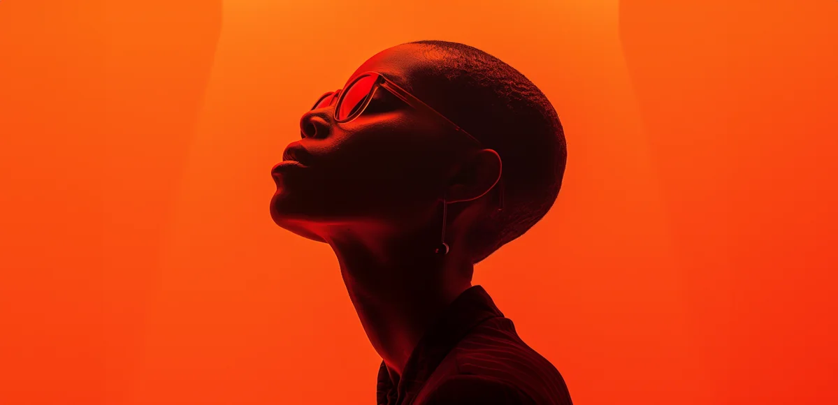 Profile of a person with short hair wearing sunglasses, against a bright orange background.