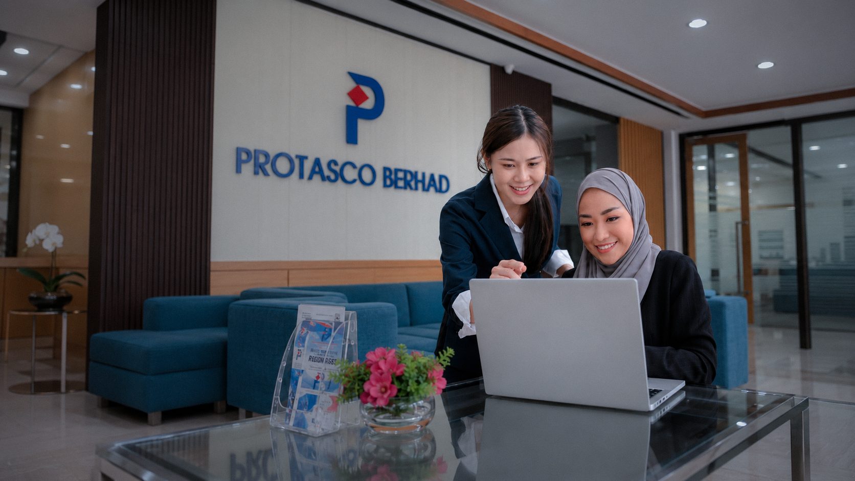 Two Protasco colleagues share a laptop in the group's lobby, beneath the Protasco Berhad sign