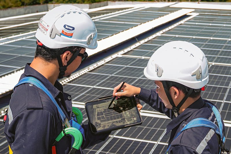 Two Protasco technicians in hard hats and full safety harnesses reviewing a tablet on a rooftop solar array