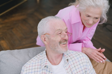 Smiling elderly couple embracing outdoors in front of green bushes on a sunny day.