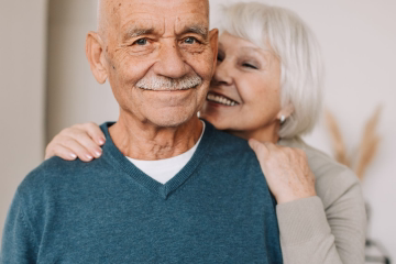 Smiling elderly couple embracing outdoors in front of green bushes on a sunny day.