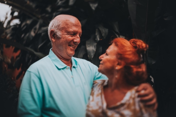 Smiling elderly couple embracing outdoors in front of green bushes on a sunny day.