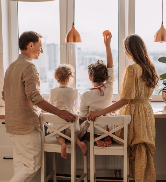 Family of four looking out a window in a kitchen, children seated on chairs and holding fruit.