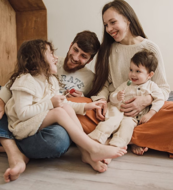 Smiling family of four sitting on floor, parents with young children holding lollipops and laughing.