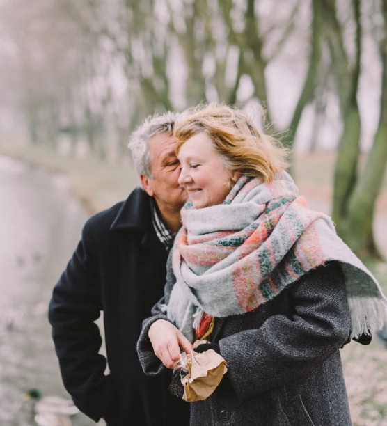 Older man giving a kiss on the cheek to a smiling woman wrapped in a colorful scarf outdoors.