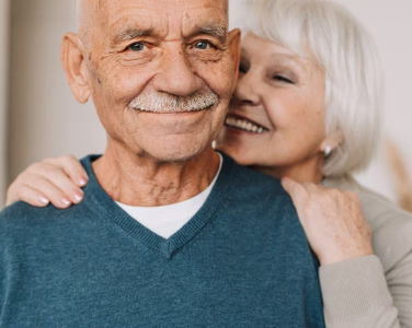 Smiling elderly man in blue sweater with woman embracing him from behind, both appearing happy.