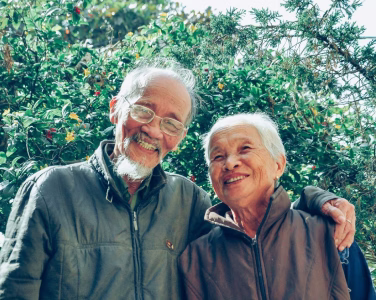 Smiling elderly couple standing close together outdoors with greenery in background.