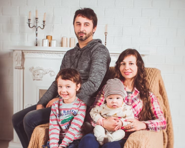 Family portrait of a man, woman, toddler, and baby seated in a cozy room with a white brick wall.