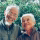 Smiling elderly couple outdoors, man in jacket with gray hair, woman with white hair and scarf.