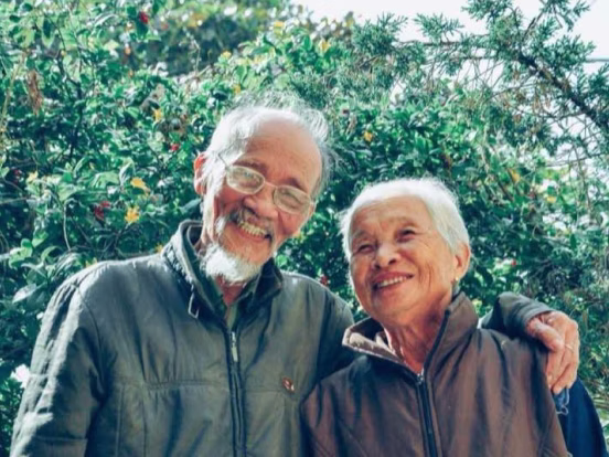 Elderly couple smiling outdoors with greenery in the background, wearing jackets and glasses.