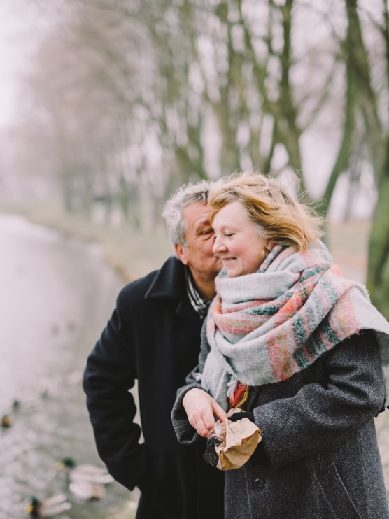 Man kissing woman on cheek as she smiles, both wearing coats and standing by a tree-lined river.