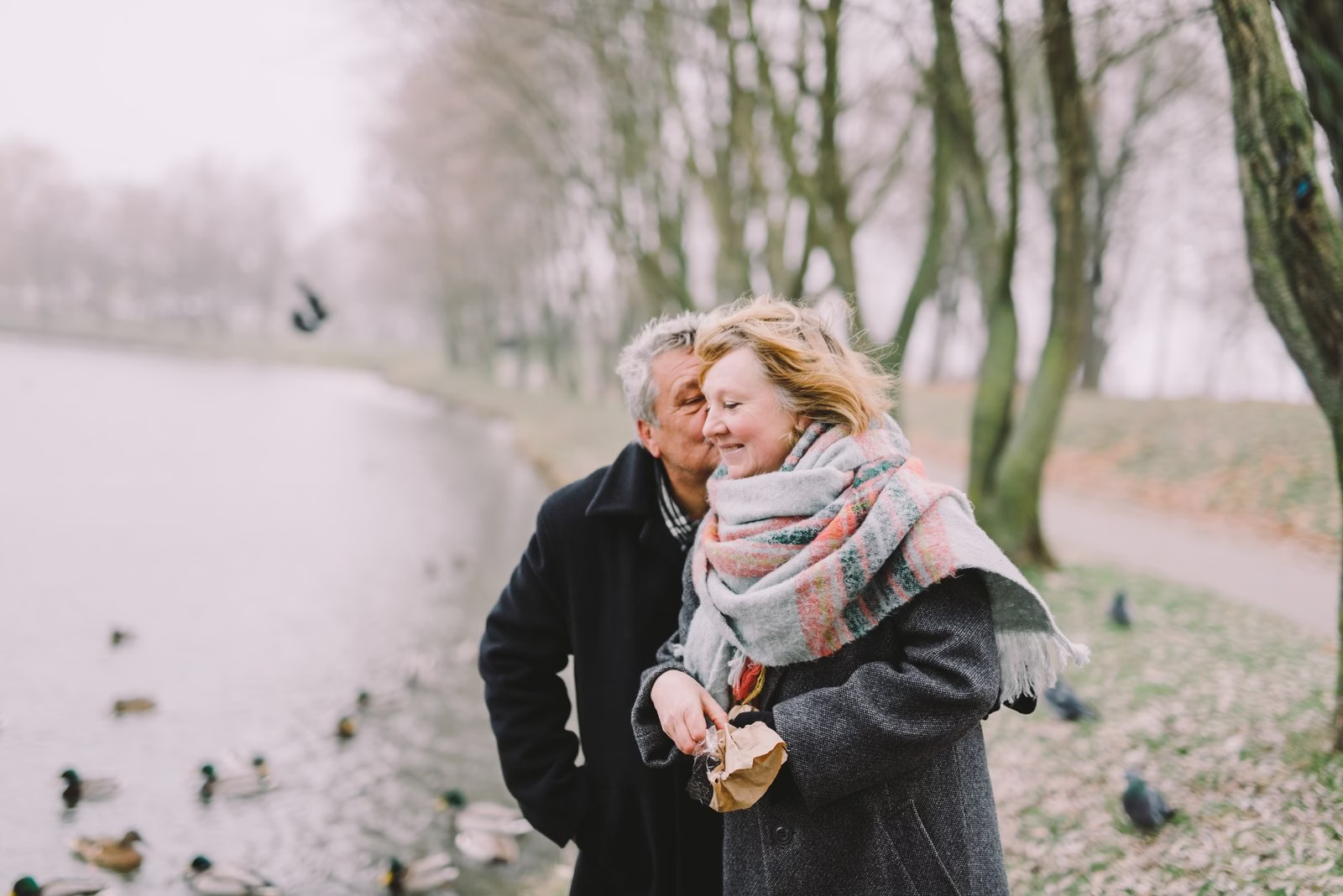 Older man kisses woman on cheek as they feed ducks by a tree-lined pond in winter.