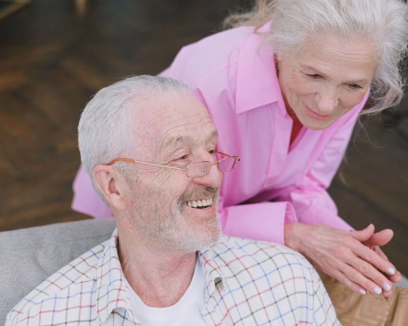 Smiling elderly man with glasses and woman in pink shirt leaning over him indoors.