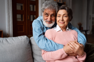 Smiling elderly couple embracing outdoors in front of green bushes on a sunny day.