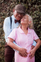 Smiling elderly couple embracing outdoors in front of green bushes on a sunny day.