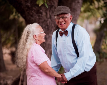Smiling elderly couple standing close together outdoors with greenery in background.