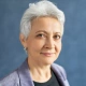 Smiling older woman with short white hair wearing a blue blazer against a plain background.