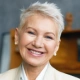 Smiling elderly woman with short white hair in a light-colored blazer indoors.