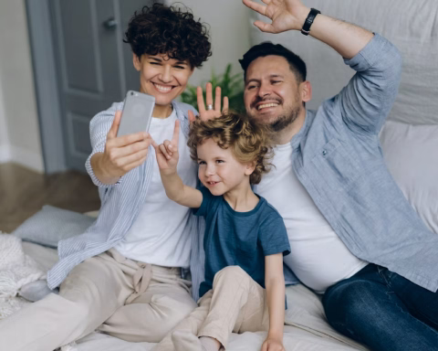 Smiling family of three sitting on bed taking selfie with smartphone and waving hands.