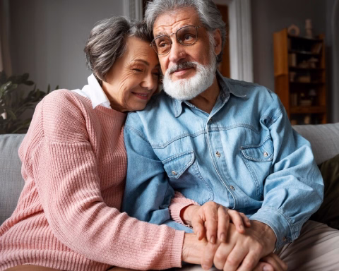 Elderly couple sitting close on a couch, woman rests head on man's shoulder, holding hands warmly.