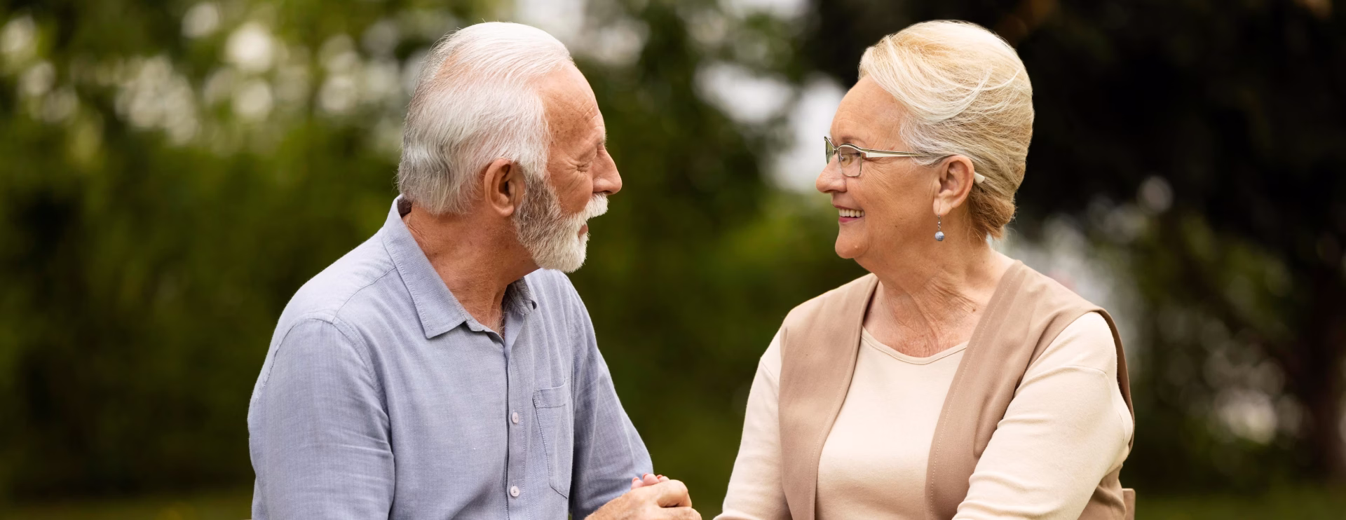 Smiling elderly couple embracing outdoors in front of green bushes on a sunny day.