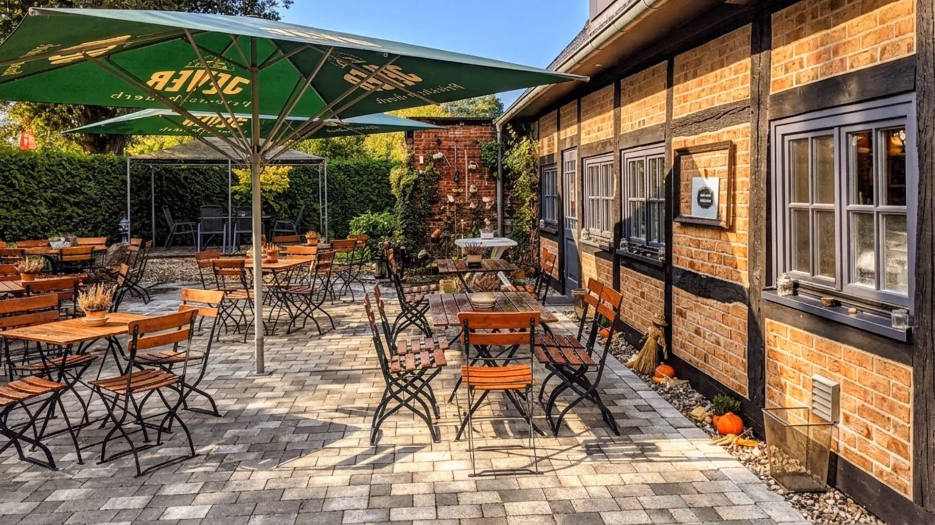 Outdoor café patio with wooden tables and chairs under a green umbrella beside a brick building.
