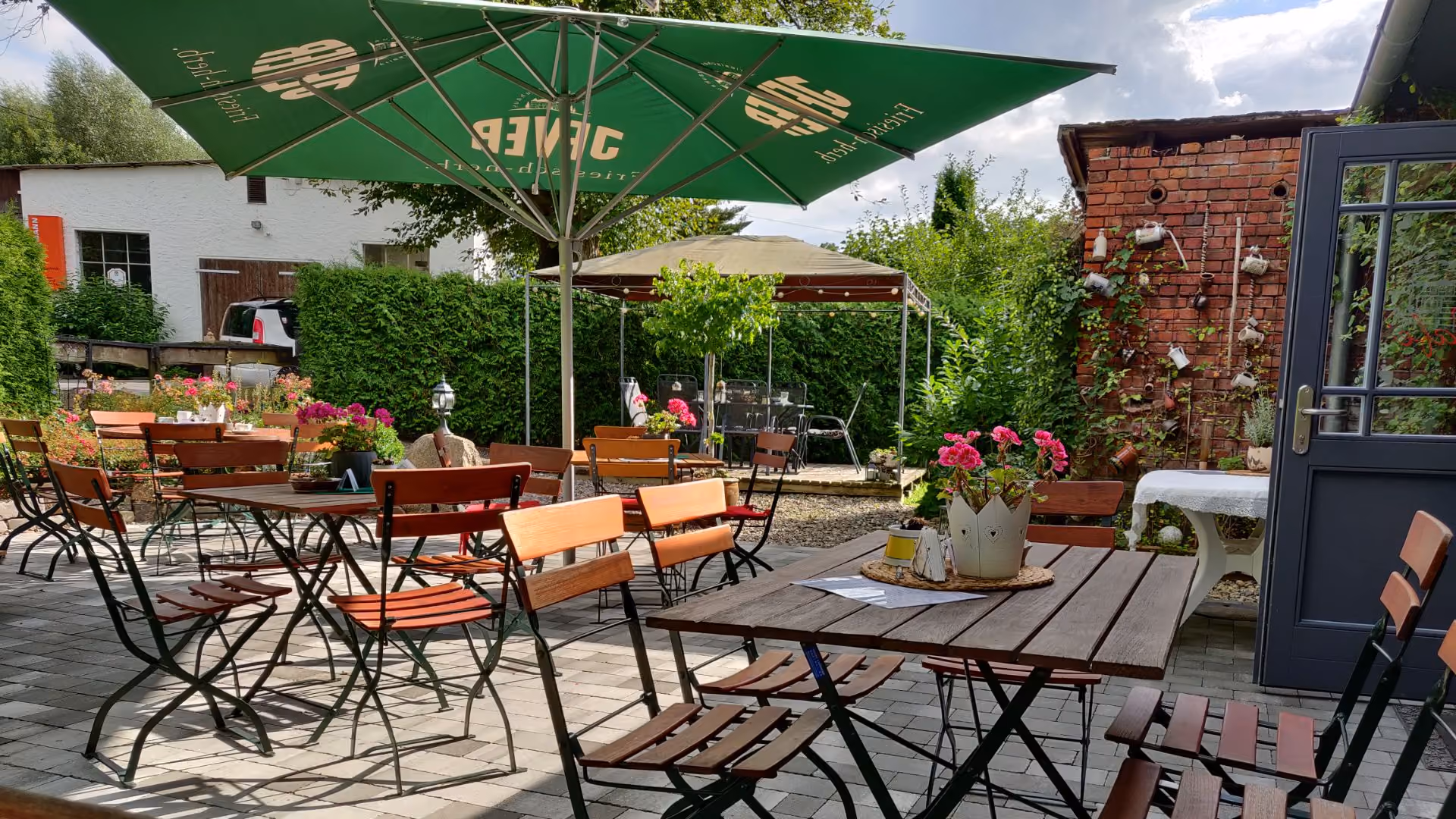 Outdoor cafe seating with wooden tables and chairs, green umbrellas, flowers, and a brick wall with hanging mugs.