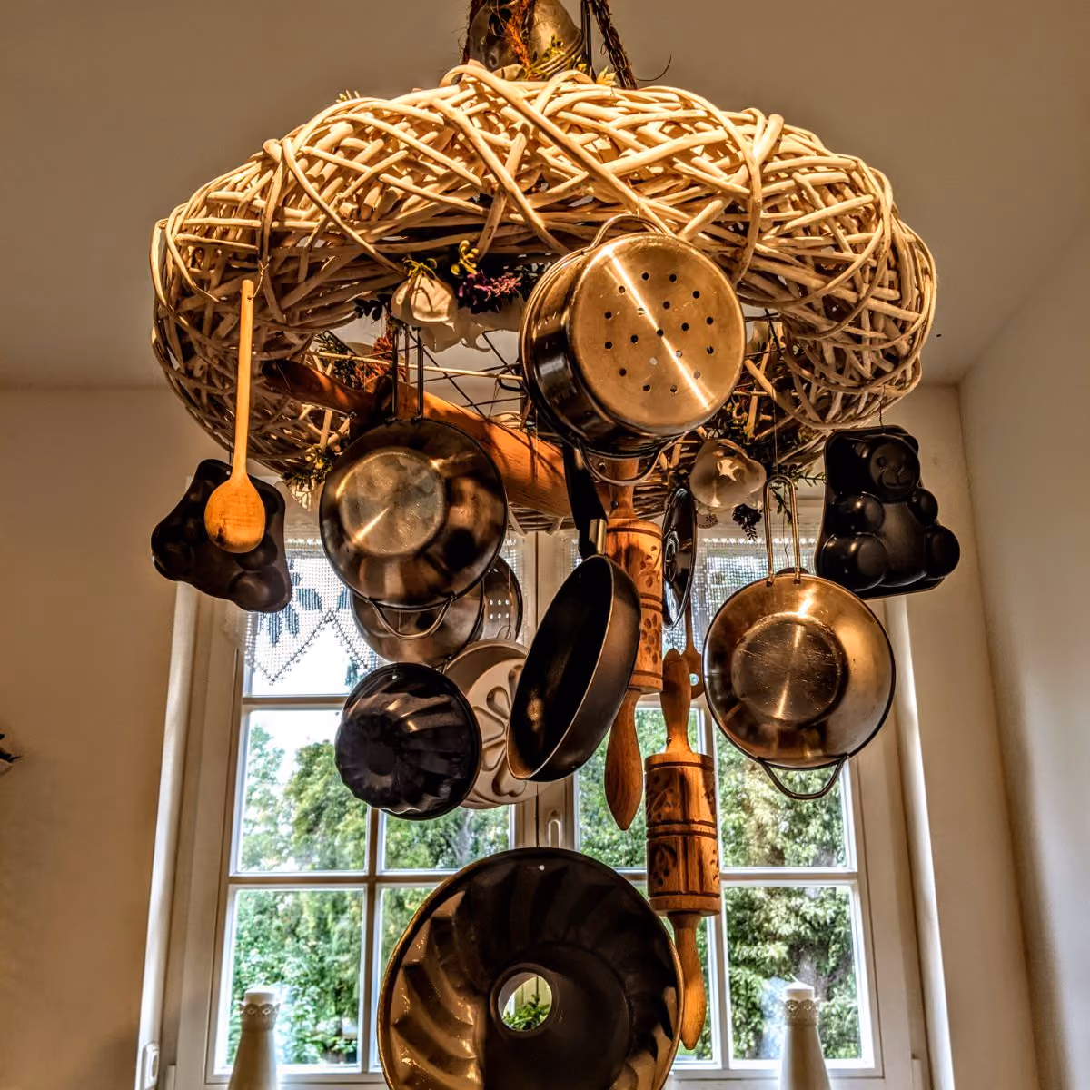 Kitchen utensil rack with pots, pans, wooden spoon, rolling pin, and baking molds hanging near a window.